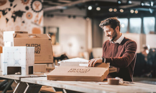 Man zit aan tafel en is een pakket aan het openen. Hij lacht vrolijk en kijkt naar het pakket.