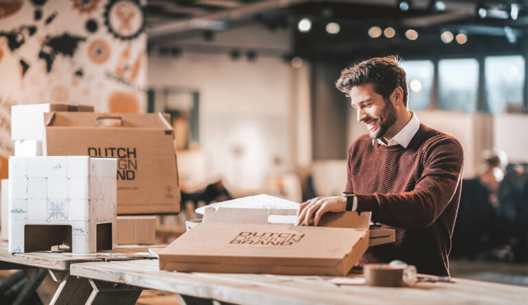 Man zit aan tafel en is een pakket aan het openen. Hij lacht vrolijk en kijkt naar het pakket.