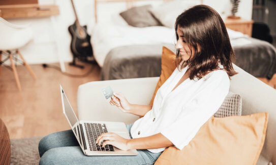 Vrouw zit achter haar laptop op de bank en gaat iets kopen. Ze heeft haar creditcard vast om te betalen.