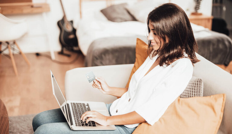 Vrouw zit achter haar laptop op de bank en gaat iets kopen. Ze heeft haar creditcard vast om te betalen.